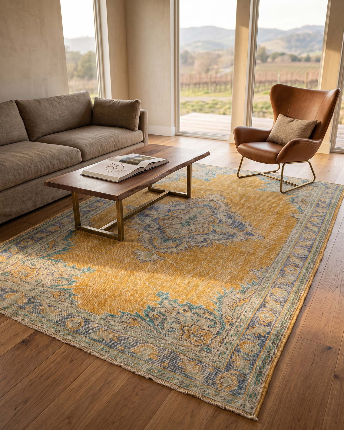 A large traditional-style area rug in mustard yellow, blue, and beige tones, styled in a modern living room with a brown leather sofa, a wooden coffee table, and a brown armchair near large windows overlooking a vineyard.