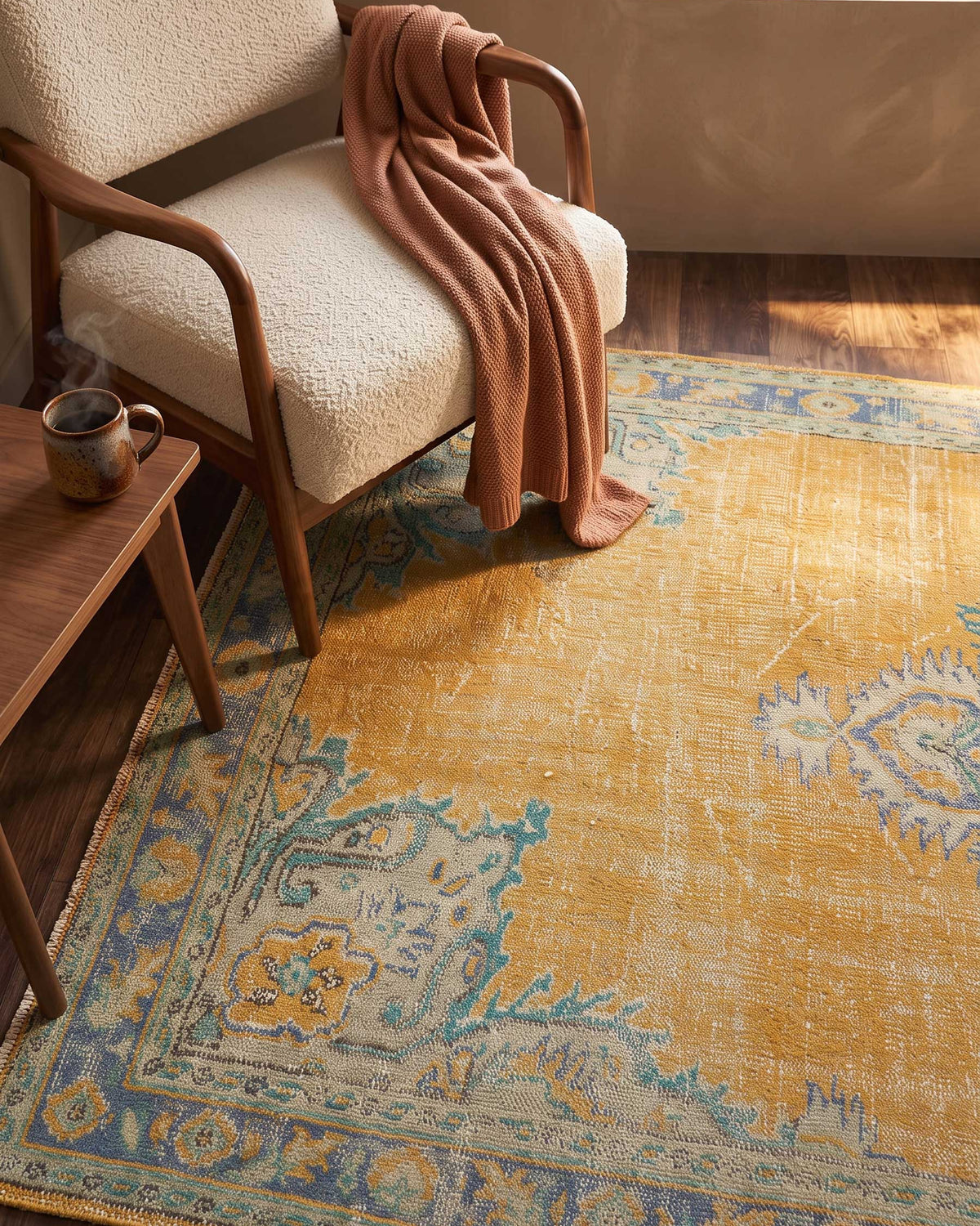 A styled interior scene showing a large vintage-style area rug in mustard yellow, blue, and beige tones, with a cream boucle armchair, a rust-colored throw blanket, and a small wooden side table with a mug.