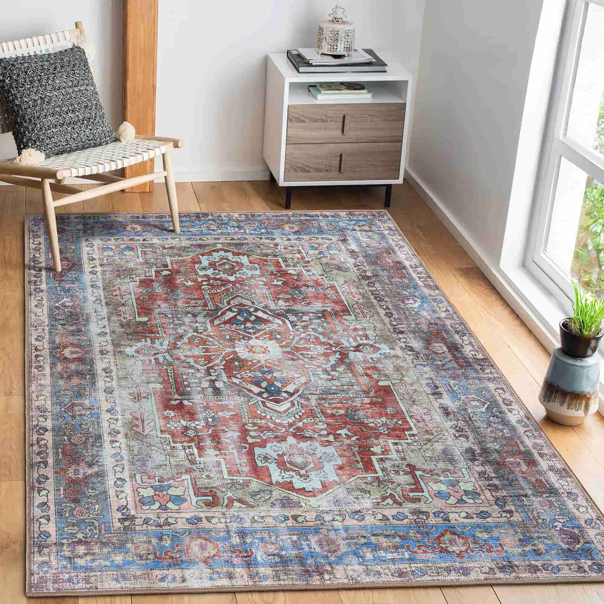 A rectangular vintage-style area rug with an intricate medallion pattern in muted red, blue, beige, and gray tones, styled on a light wood floor in a bright room next to a chair and a side table.