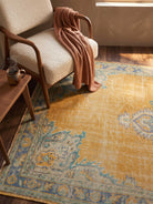 A styled interior scene showing a large vintage-style area rug in mustard yellow, blue, and beige tones, with a cream boucle armchair, a rust-colored throw blanket, and a small wooden side table with a mug.