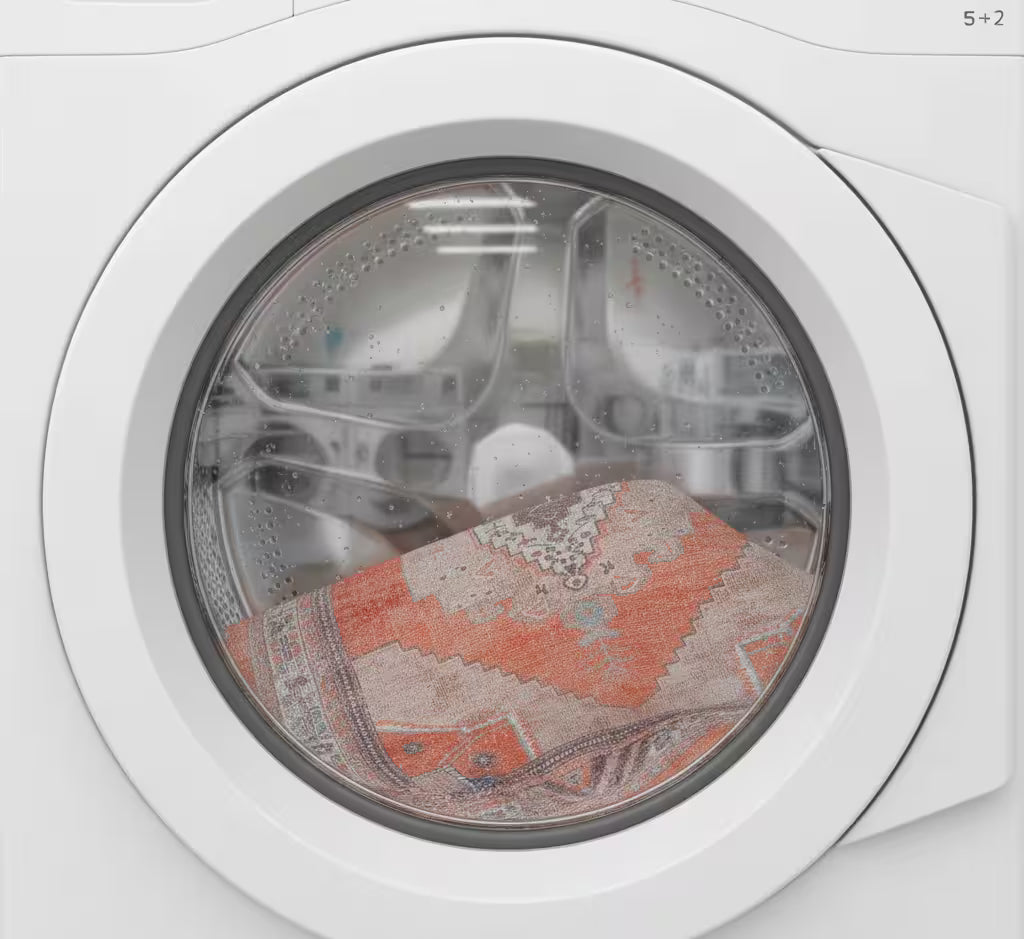 A close-up of a white front-loading washing machine door with a clear circular window, showing a patterned orange and beige rug or textile inside the drum. Fame Rugs.