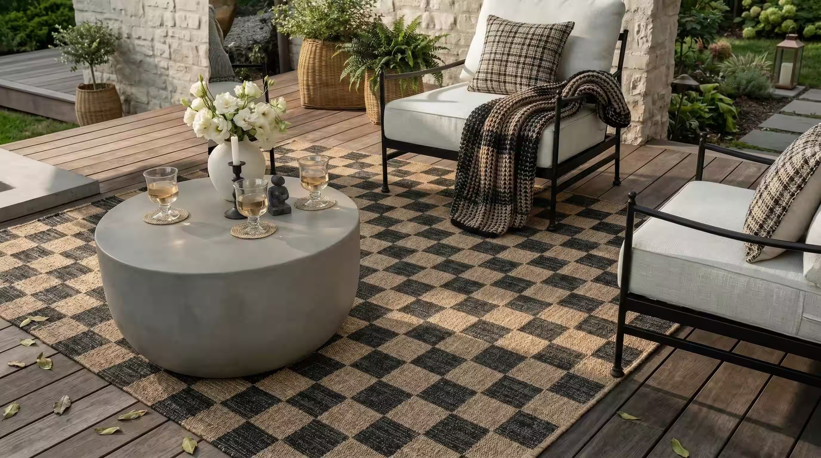 Outdoor patio furniture set on a checkered black and beige rug, featuring a round concrete coffee table, white cushioned armchairs with plaid pillows, and a throw blanket on a wooden deck. Fame Rugs.
